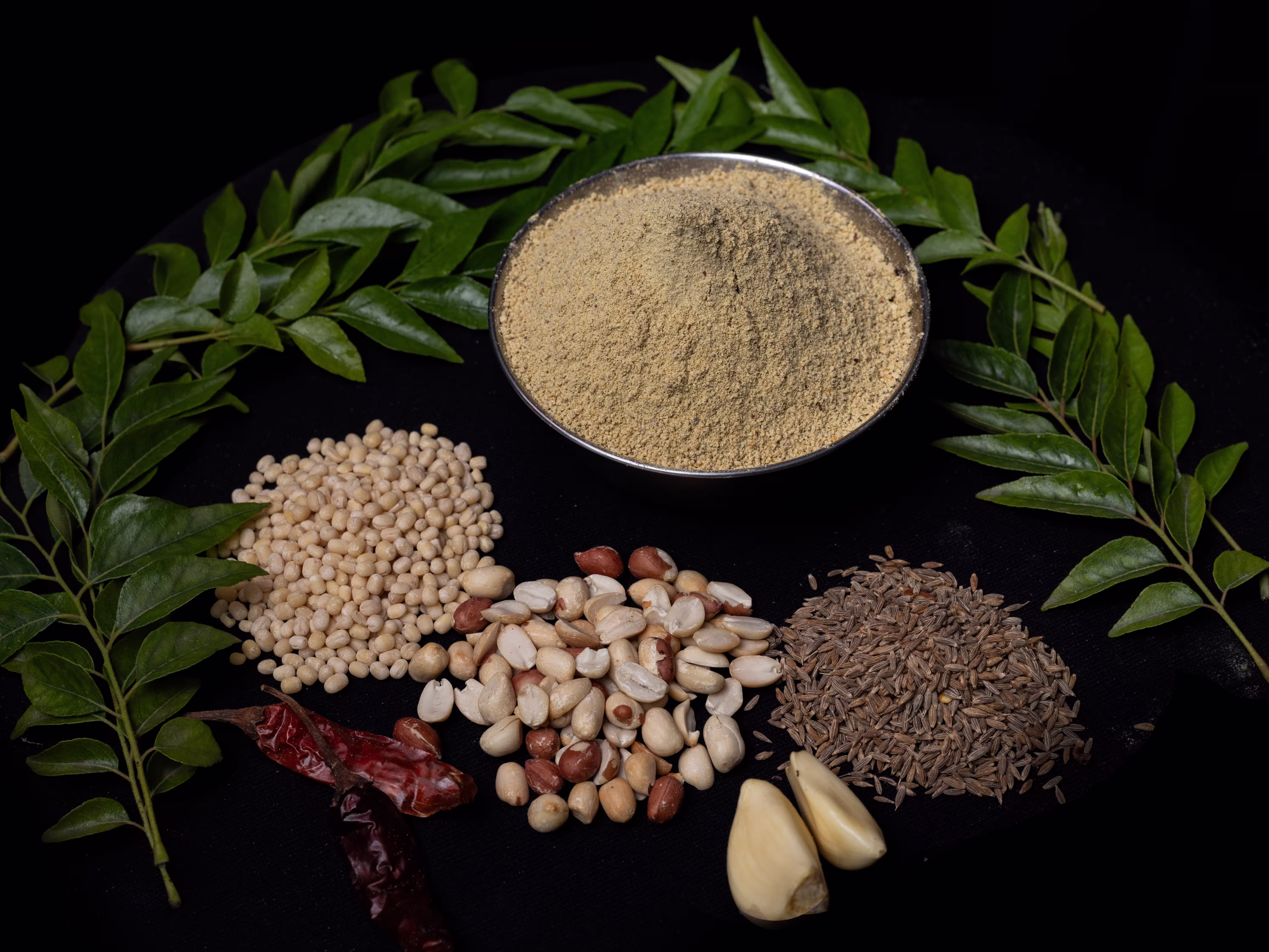 Curry leaves against a black background with red chilies, nuts, and a bowl of spice.